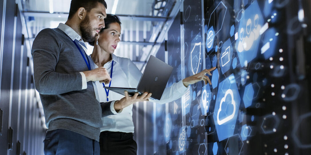 Male IT Specialist Holds Laptop and Discusses Work with Female Server Technician. They're Standing in Data Center, Rack Server Cabinet with Cloud Server Icon and Visualization.