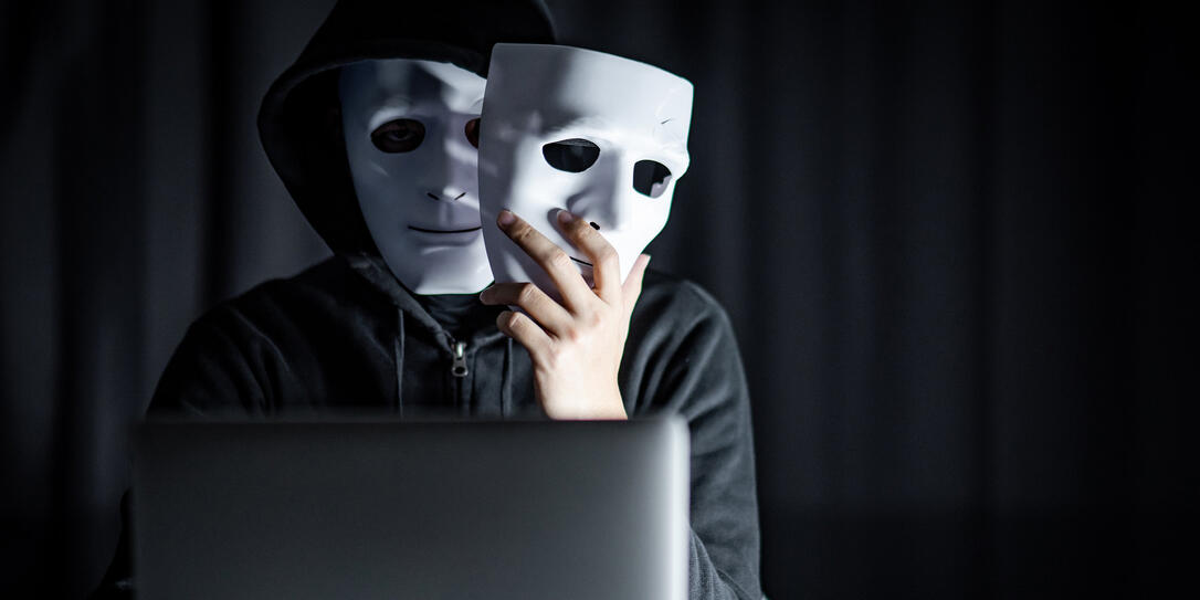 Mystery male hoodie hacker wearing mask holding white mask sitting with laptop computer on the table. Anonymous social masking. Ransomware cyber attack or internet security concepts