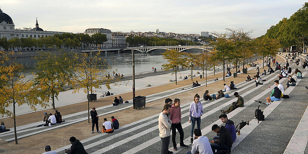 Menschen sitzen entlang des Flusses Rhone. Die französischen Behörden haben die Stadt Lyon auf die höchste Warnstufe gesetzt. Versammlungen sind untersagt und Bars werden schließen müssen. Foto: Laurent Cipriani/AP/dpa