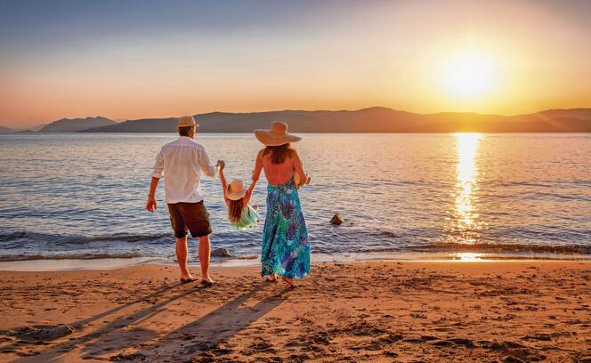 A family on holidays stands on a beach in the mediterranean sea and enjoys the summer sunset time