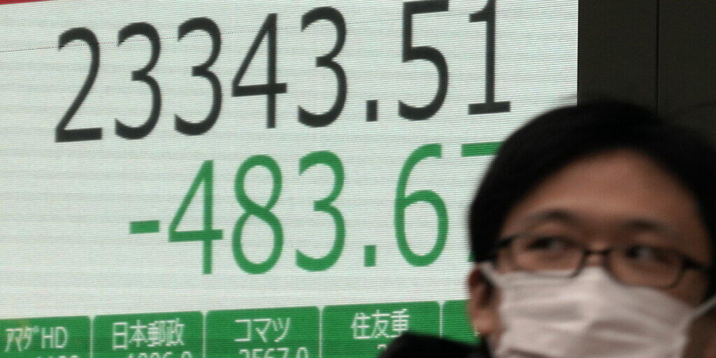 An den Börsen geht es wegen dem Coronavirus abwärts: ein Mann mit einer Maske in Tokio vor einer Börsenanzeigetafel.